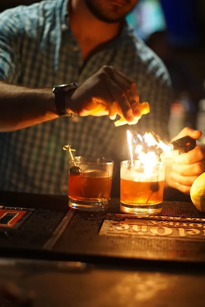 Bartender flaming a drink with citrus peel in a dimly lit bar.