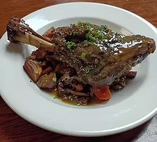 Braised lamb shank with roasted vegetables and herbs on a white plate.