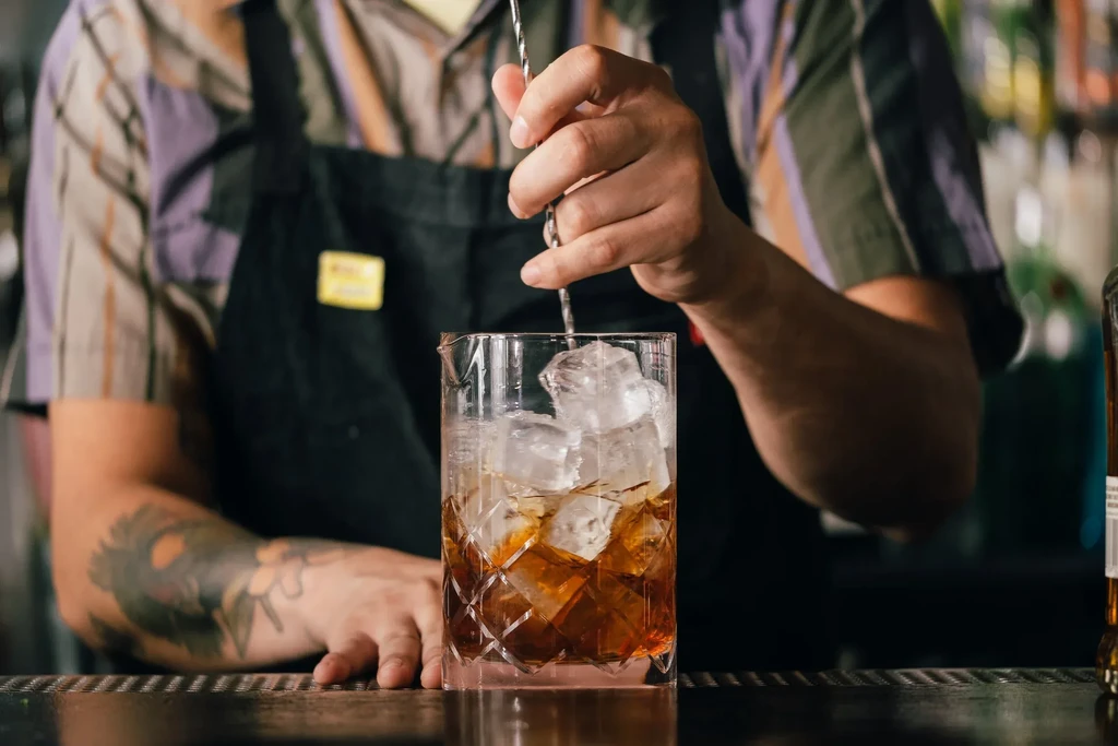 Bartender's hands stir an amber cocktail in a crystal glass with ice.
