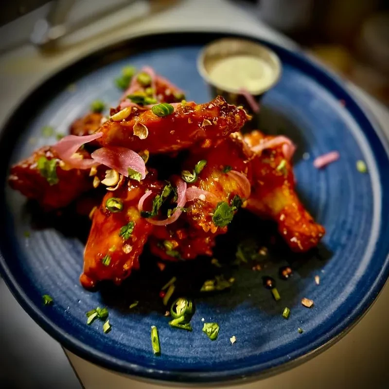 Chicken wings with reddish-orange sauce, pickled onions, scallions, and peanuts.