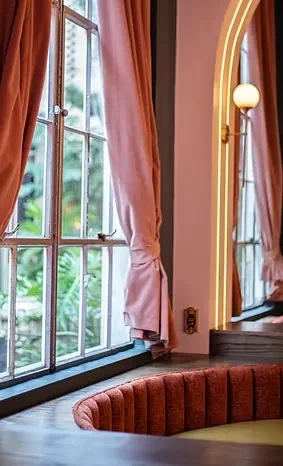 Restaurant interior with window, peach velvet curtains, arched mirror, and orange banquette.