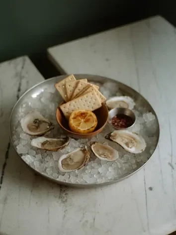 Close-up of six oysters on ice with crackers, lemon, and sauce.
