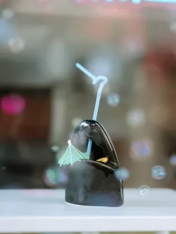 Misty drink in dark, creature-shaped vessel with straw, umbrella, bubbles.