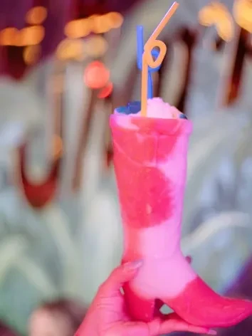 Frothy, layered drink in boot glass, held in dim pink light.