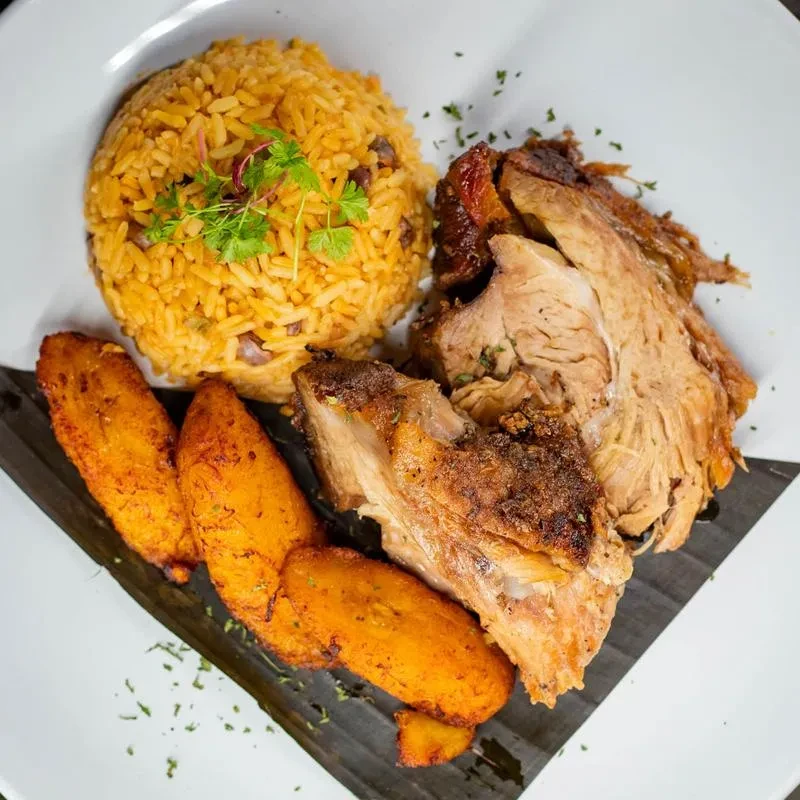 Seasoned yellow rice, roasted meat, and fried plantains on a dark leaf.
