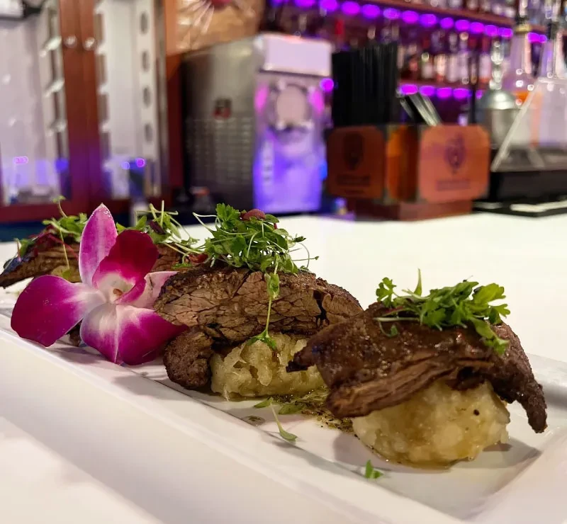 Appetizer: Sliced seared meat, microgreens, orchid, blurred purple-lit bar.