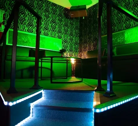 Green-lit lounge with ornate wallpaper, raised seating, and blue-lit steps.
