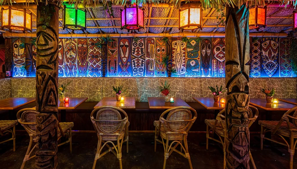 Exotic tiki bar interior with carved pillars, colorful masks, and glowing lanterns.