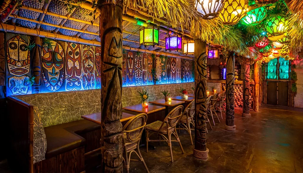 Tiki bar interior with carved masks, totems, thatched roof, and lanterns.