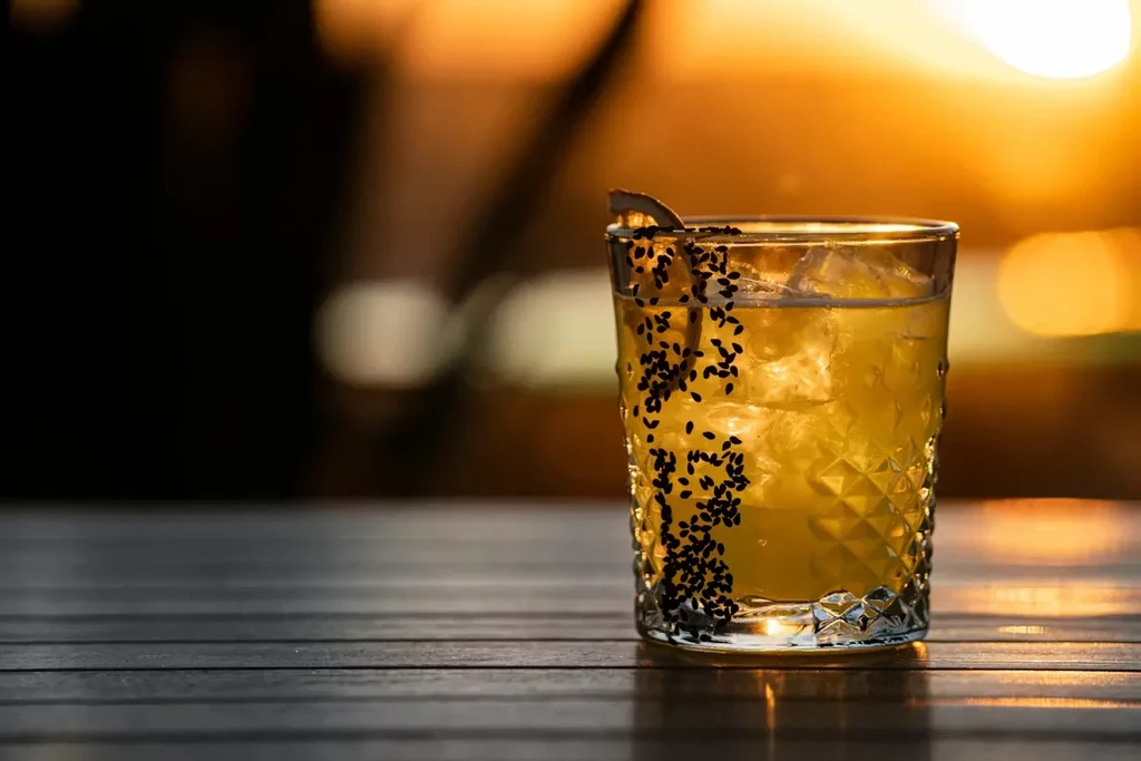 Clear yellow cocktail with sesame seeds on textured glass rim, warm bokeh.