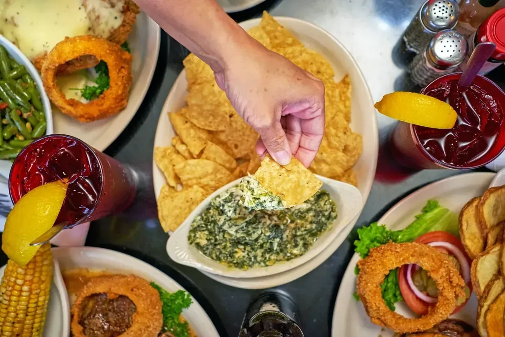 Overhead view: hand dips chip into spinach dip amidst comfort food.