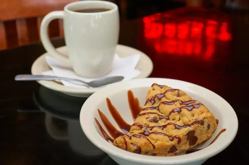 Chocolate chip cookie slice with chocolate drizzle, coffee mug on dark table.