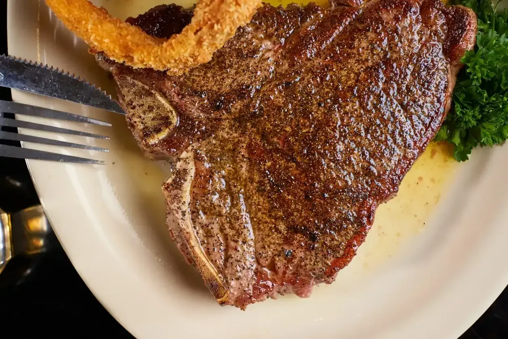Juicy seared T-bone steak, onion ring, parsley, and cutlery on plate.