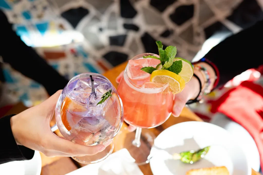 Two hands holding two colorful, garnished cocktails.