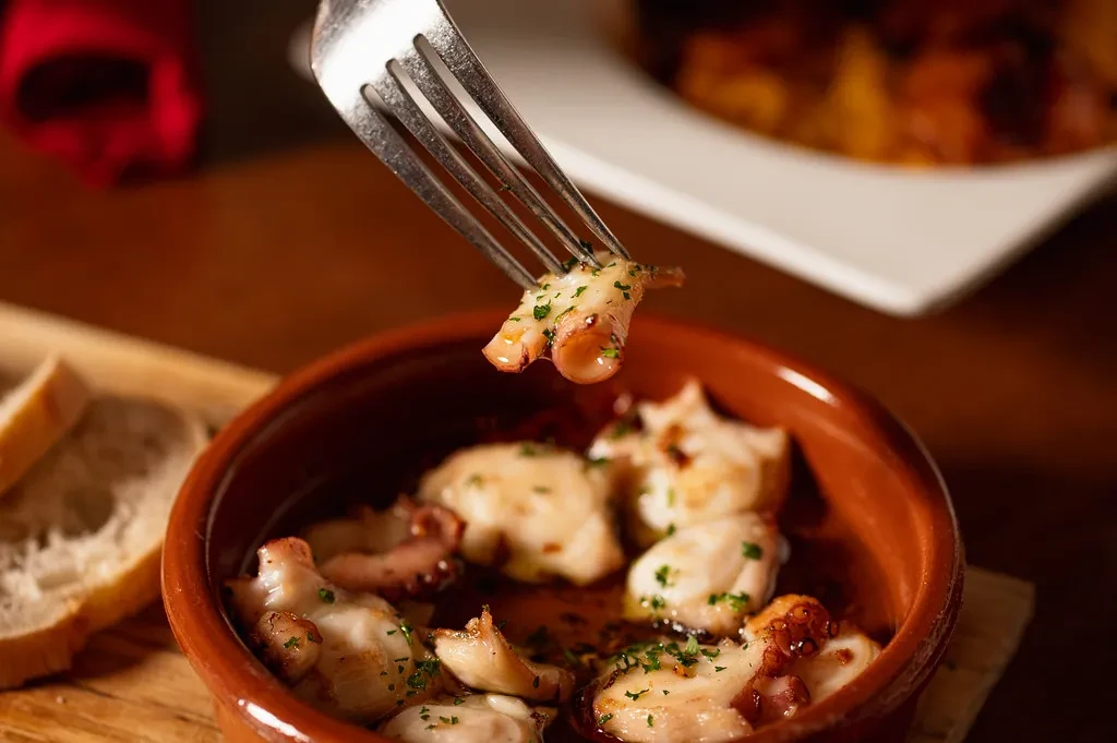 Close-up of fork lifting purple-white octopus from rustic terracotta bowl.