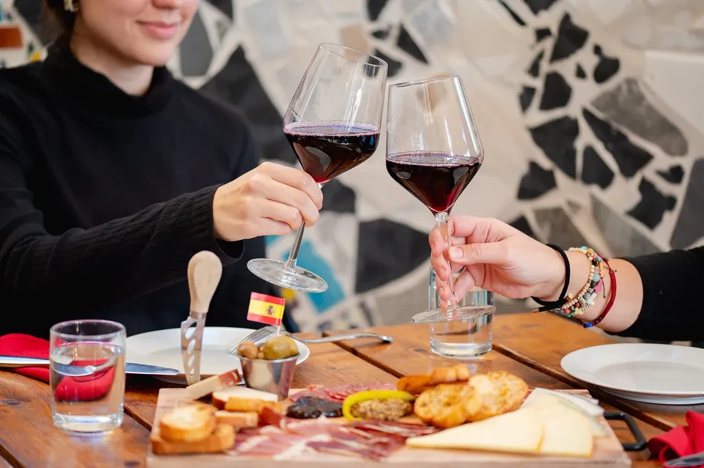 Two people toast red wine over a large charcuterie board.