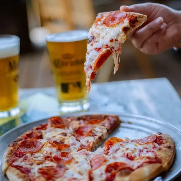 Hand lifts a pepperoni pizza slice with cheese pull, two beers.