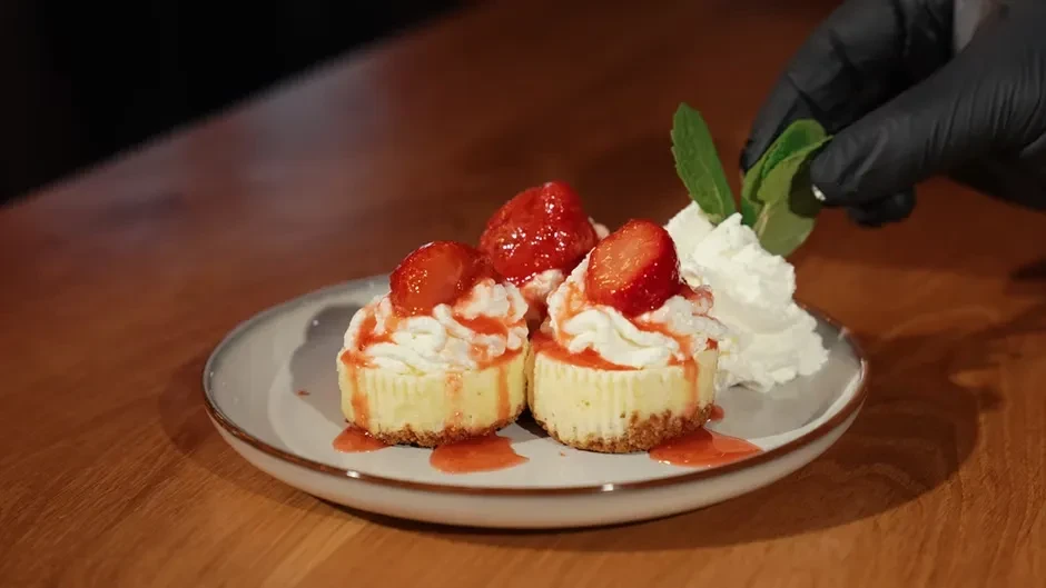 Gloved hand garnishes three strawberry desserts with whipped cream and mint.