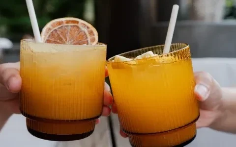 Two hands holding textured amber glasses of orange drinks with garnishes.