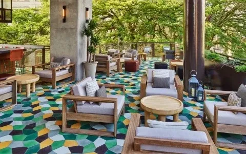Empty outdoor lounge area with mosaic floor, wooden furniture, and lush greenery.