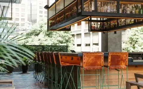 Outdoor urban bar with wooden counter, leather stools, industrial shelving.