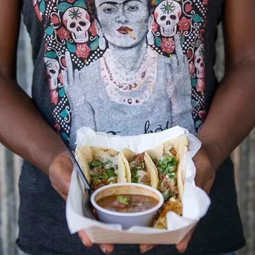 Person in Frida Kahlo t-shirt holds three tacos and dipping sauce.