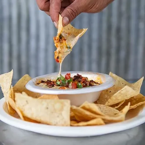 Hand dipping chip into gooey cheese dip with pico and meat.