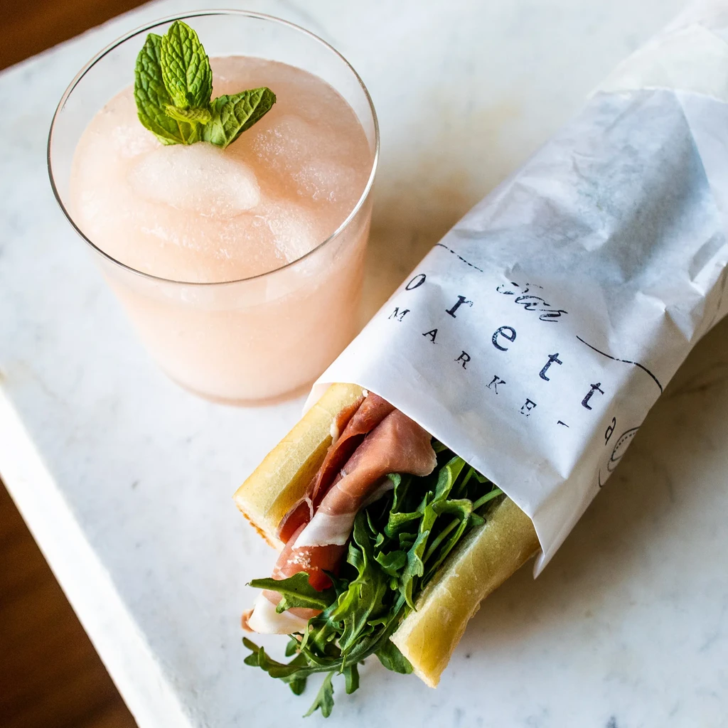 Overhead: Pink frosty drink with mint, branded prosciutto and arugula sub.