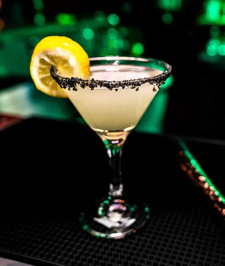Cocktail in martini glass, dark rim, lemon garnish, on black textured mat.