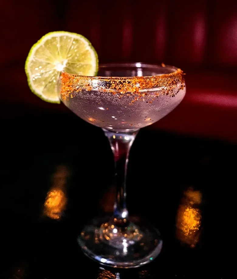 Light cocktail in frosty coupe glass with a seasoned rim and lime.