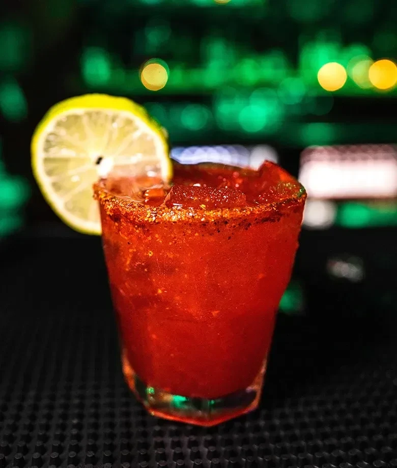 Red cocktail in faceted glass with chili-salt rim and lime.