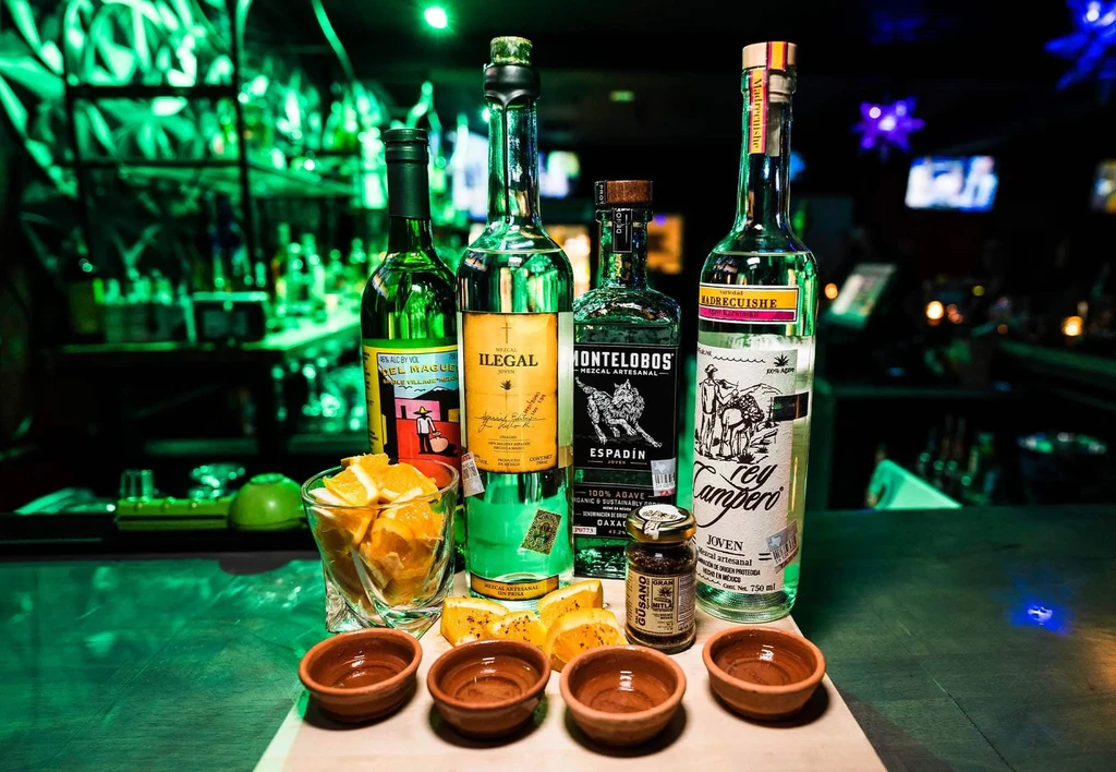 Four mezcal bottles, orange slices, and ceramic cups on a bar.