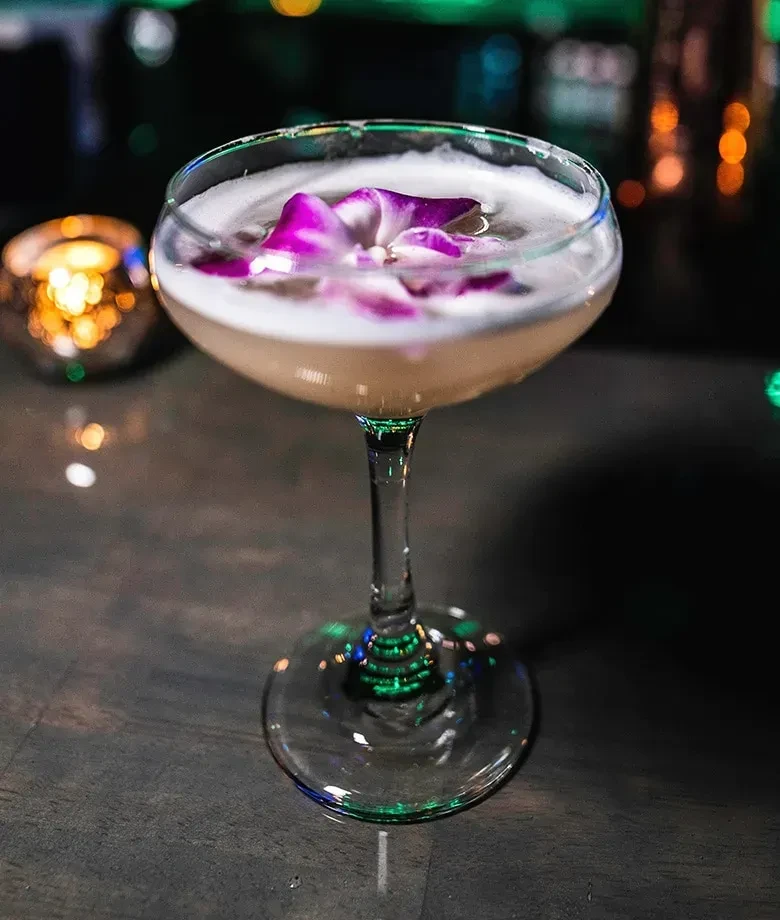 Creamy cocktail with purple orchid petals in a coupe glass.