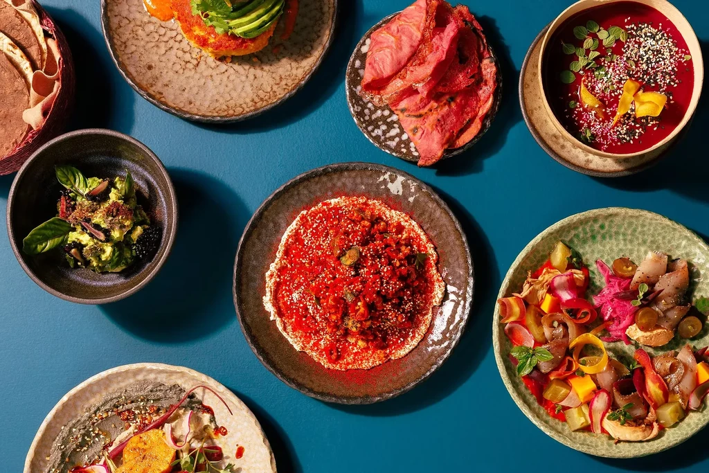Overhead shot of seven colorful artisanal dishes on a teal surface.