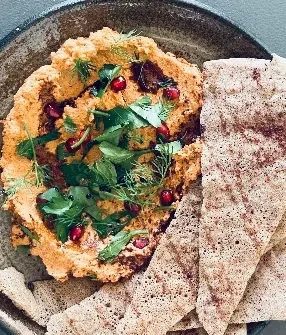 Thick orange dip with herbs, pomegranate, and flatbread in dark bowl.