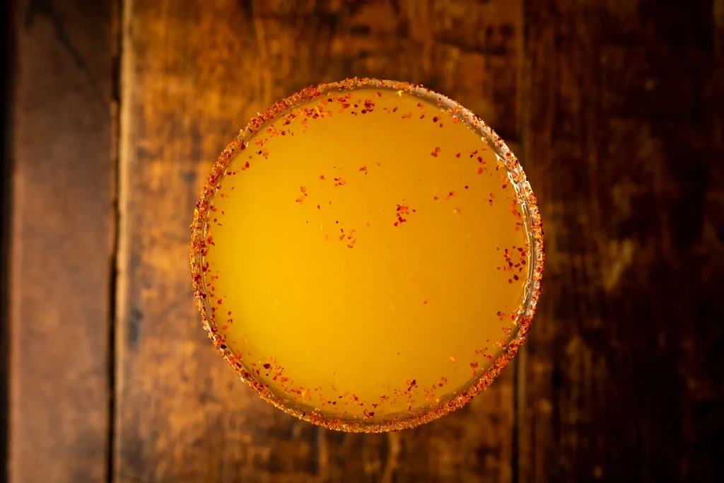 Yellow-orange cocktail with a chili-spiced rim, overhead on a wooden table.