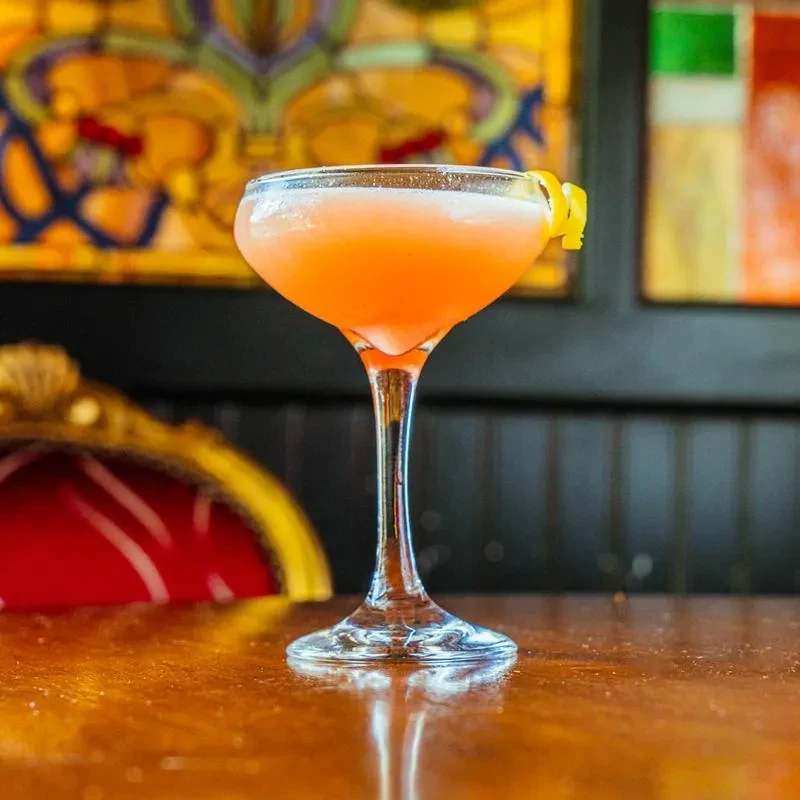Orange-pink cocktail with lemon peel twist and stained-glass background.