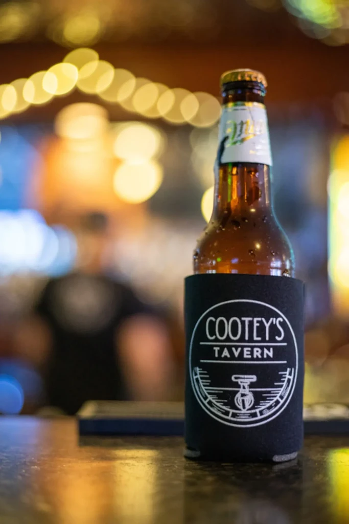 Cold brown beer bottle in Cootey's Tavern koozie on a bar top.