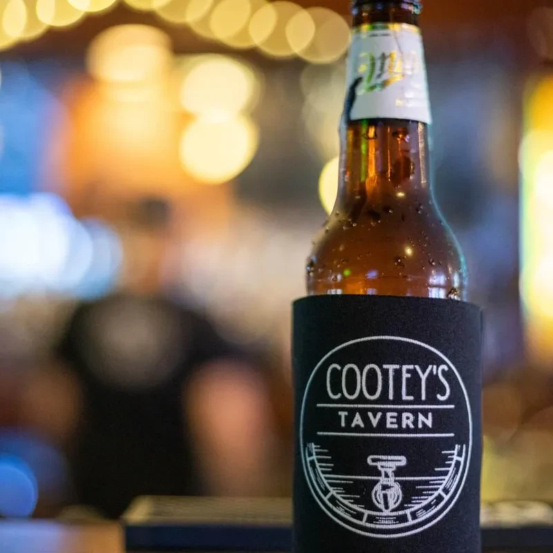 Amber beer bottle with condensation in a 'COOTEY'S TAVERN' koozie.