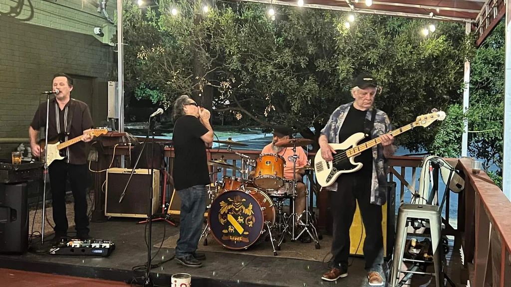 Four men play guitar, harmonica, drums, and bass on an outdoor stage.