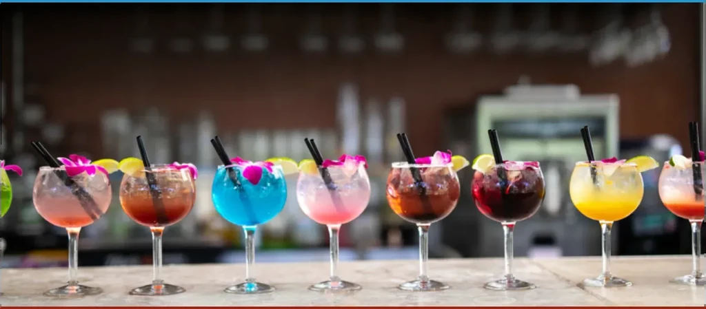 Eight colorful, garnished cocktails in stemmed glasses on a bar.