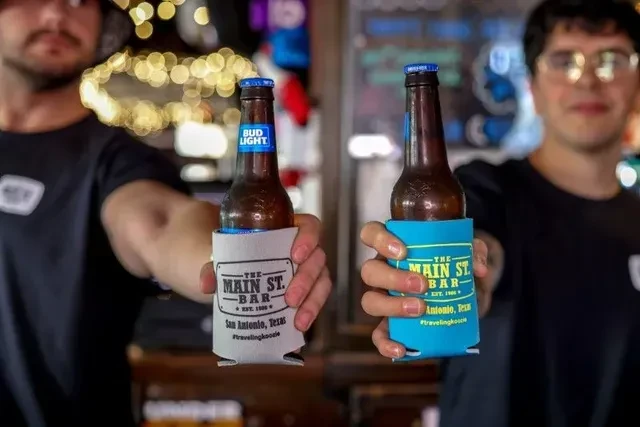 Two people hold beers in koozies with 'THE MAIN ST. BAR' logo.