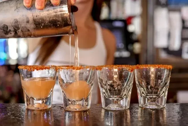 Hands pouring orange liquid into seasoned shot glasses at a bar.