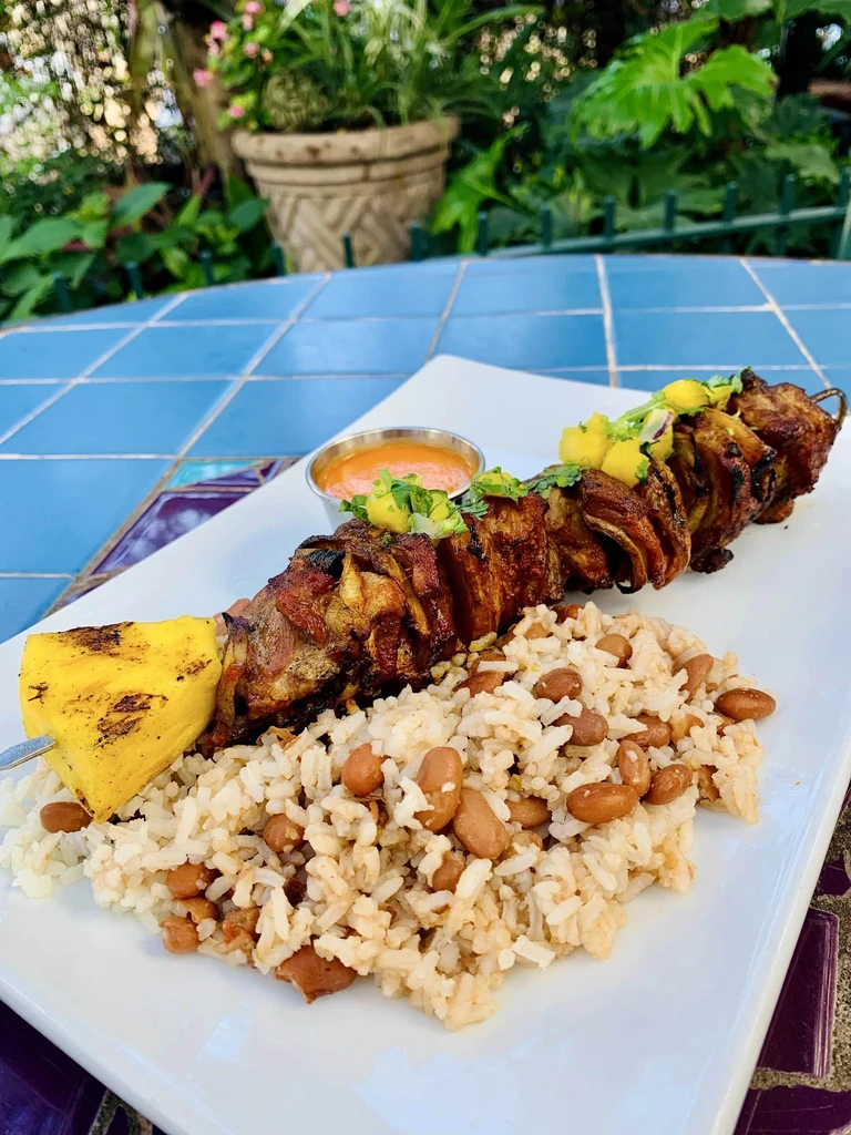Grilled meat skewer, pineapple, rice, beans, salsa on blue tiles.