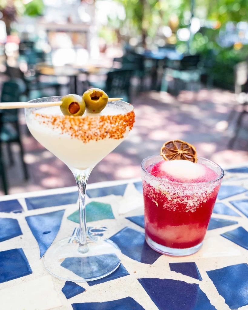Two garnished cocktails, red and white, on an outdoor mosaic table.