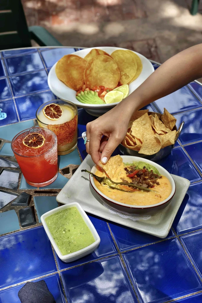 Overhead view of hand dipping chip into queso, food, and cocktails.