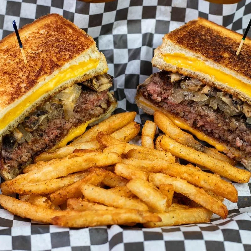 Close-up of a patty melt, seasoned fries, on black and white checkered paper.