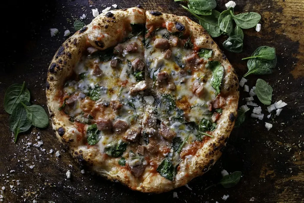 Overhead wood-fired pizza with meat, spinach, and melted cheese.