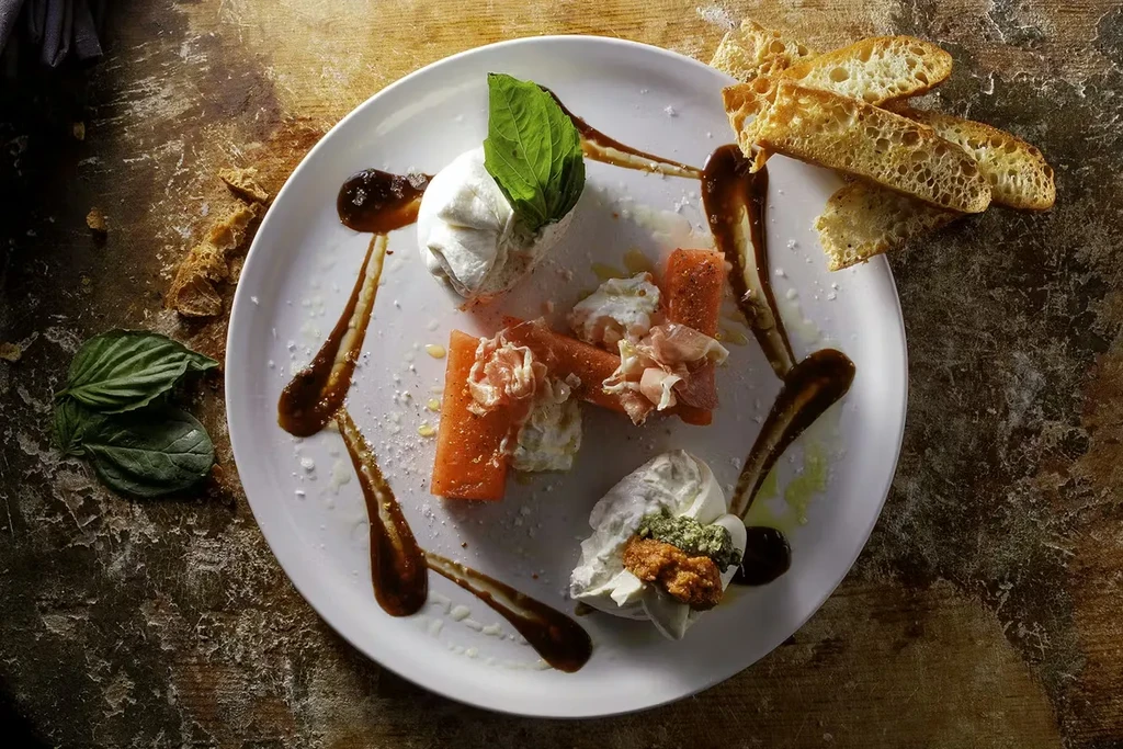 Overhead view of burrata, prosciutto, melon, and toasted bread appetizer.