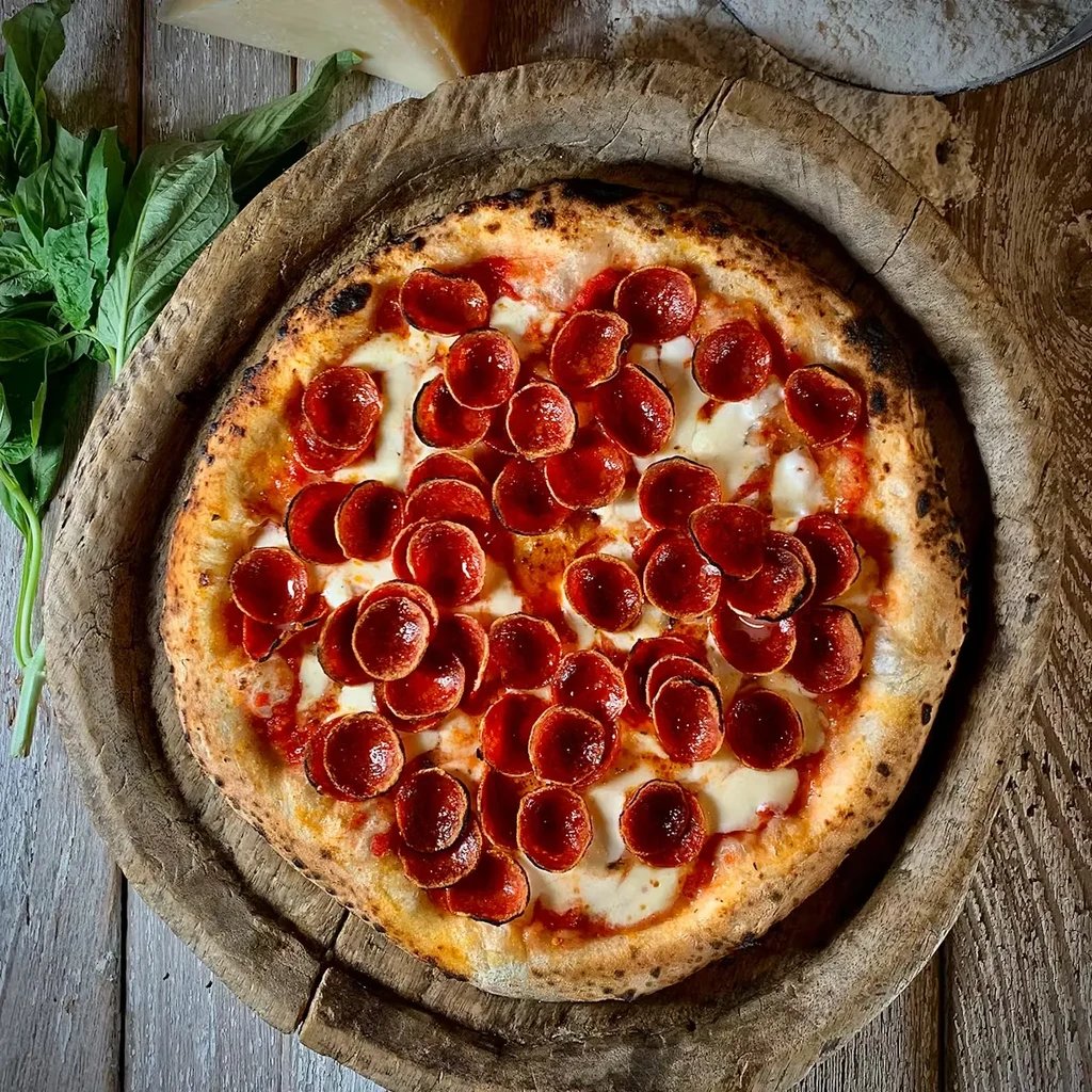 Overhead: Neapolitan pepperoni pizza, charred crust, fresh basil on wood board.